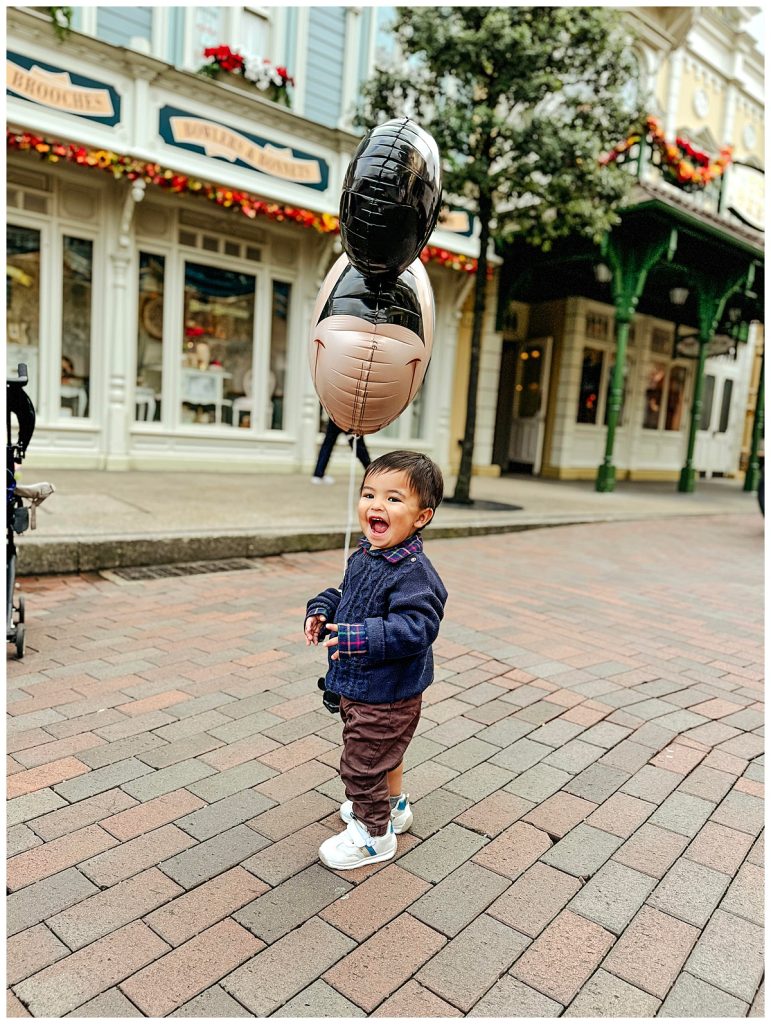 A happy toddler holding a Mickey Mouse balloon, laughing and playing on Main Street at Disneyland Paris. The child is dressed warmly in a navy sweater and brown pants, with the iconic Disney storefronts and festive decorations visible in the background. The photo captures the magical experience and joy of visiting Disneyland Paris with young children.
