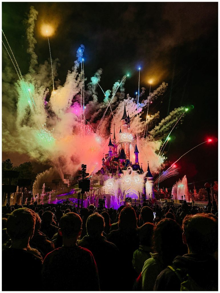 Spectacular fireworks display lighting up the night sky above Sleeping Beauty Castle at Disneyland Paris, with a crowd of onlookers enjoying the colorful show. The scene is filled with vibrant colors and magical effects, highlighting the iconic castle and creating a breathtaking moment